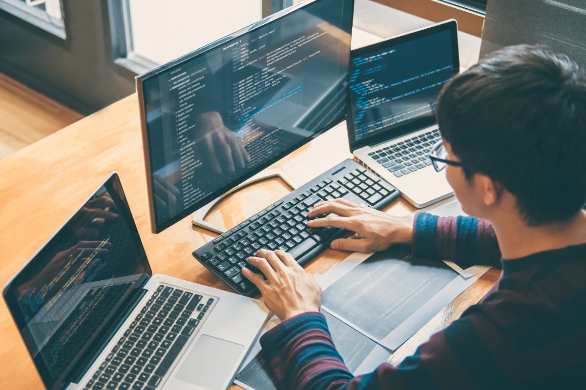 Man with glasses sitting infront of 2 laptions and one computer screen typing on a keyboard.