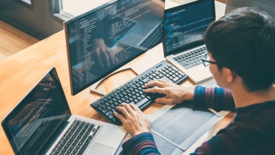 Man with glasses sitting infront of 2 laptions and one computer screen typing on a keyboard.